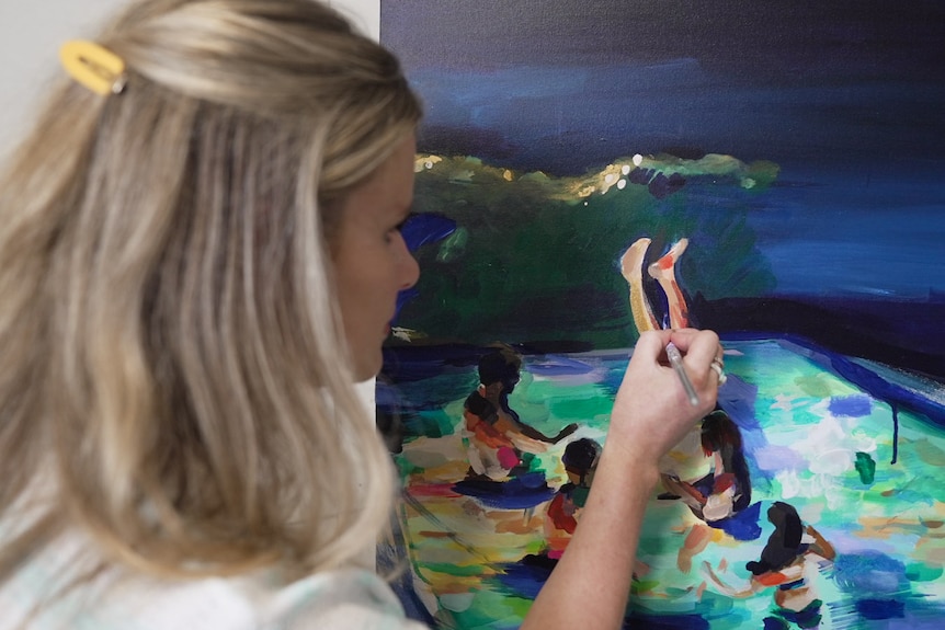 Side profile of Bronte, up close, painting a dark blue sky, painting shows people in a pool underneath the sky, some legs up.