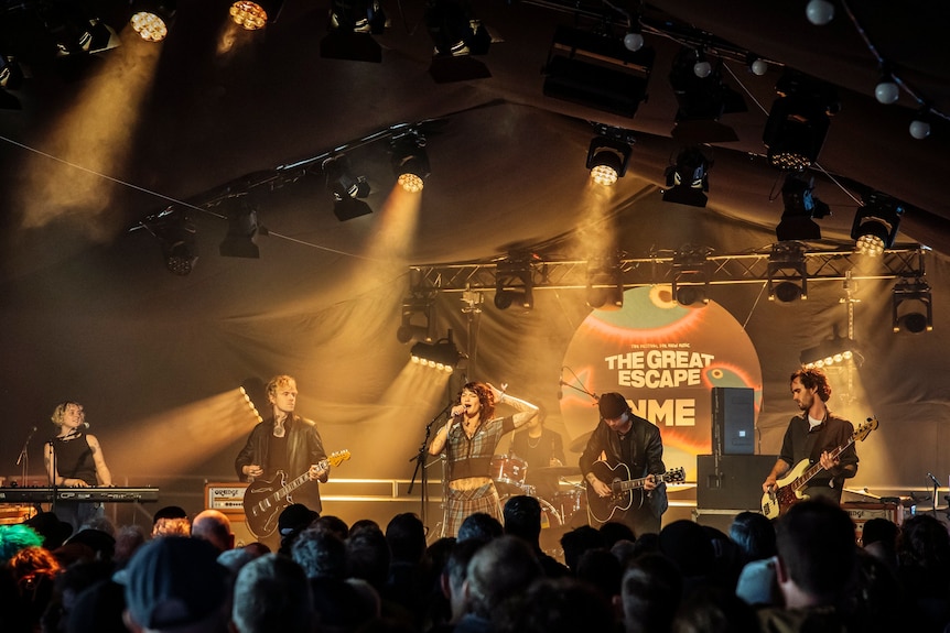 A band playing music on-stage as the lead singer holds a microphone, a sign in the background reads: THE GREAT ESCAPE