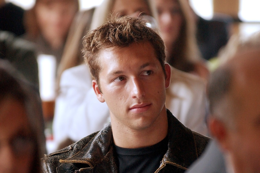 Ian Thorpe sits in a room of out-of-focus people in front and behind him. He looks off to the side thoughtfully.