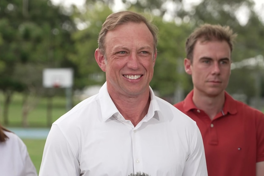 A man in a white button shirt smiling with another man in orange polo behind him.