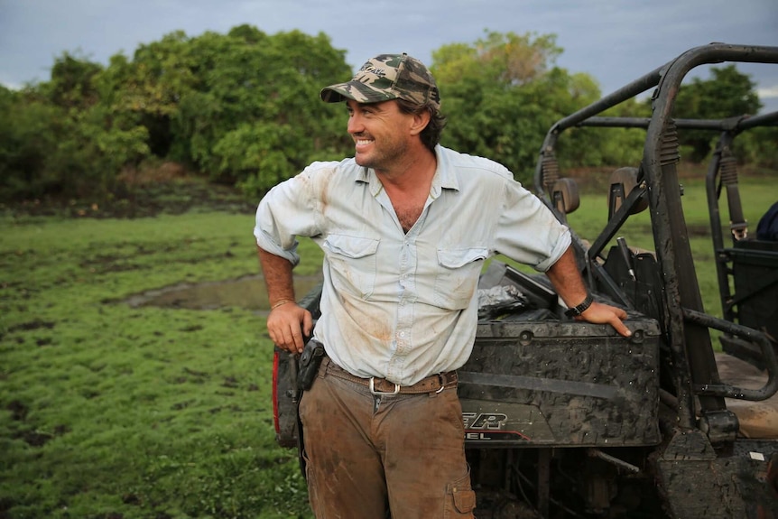 A man in work clothes, wearing a cap and leaning on against an off-road vehicle.