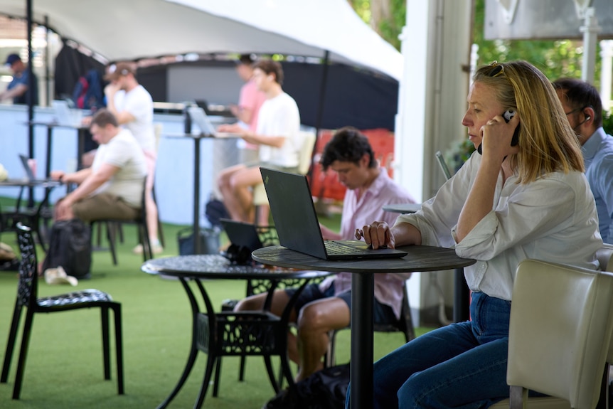 A woman talks on her phone while operating a laptop in an outdoor office area at a cricket ground.
