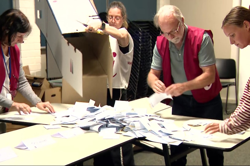 Three people in red and white vests stand at a table with their hands on leaflets as a woman pours more from a cardboard box.