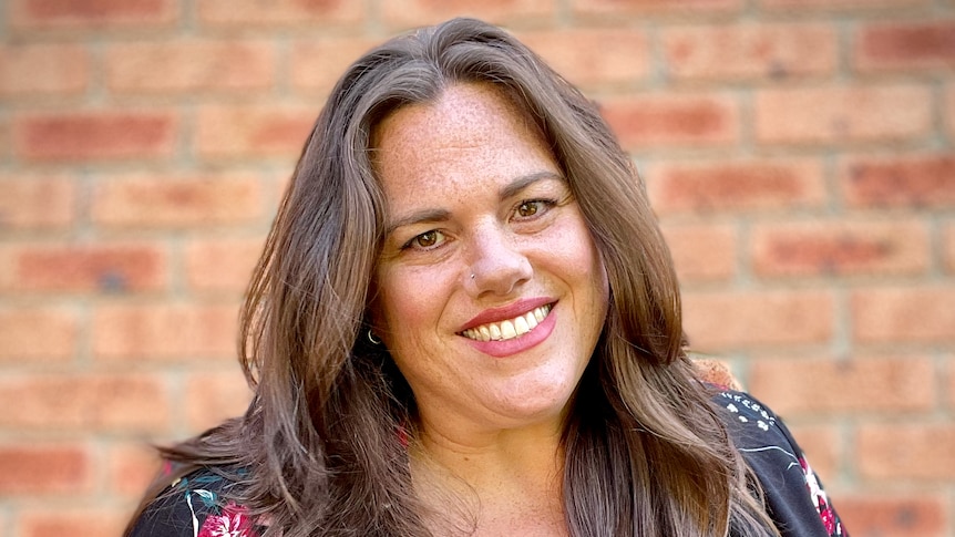 Liz Allen, wearing black floral blazer over navy top, smiles outside in front of a brick wall