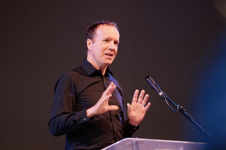 A man speaking at a lecturn in a dark shirt and with a dark background