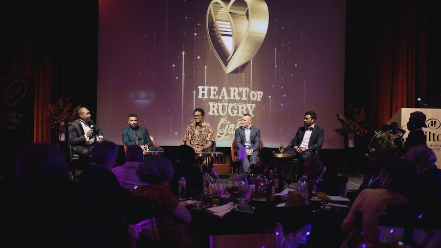 Five well known rugby players sit in chairs on a stage with a sign 'Heart of Rugby gala' in the background on a screen