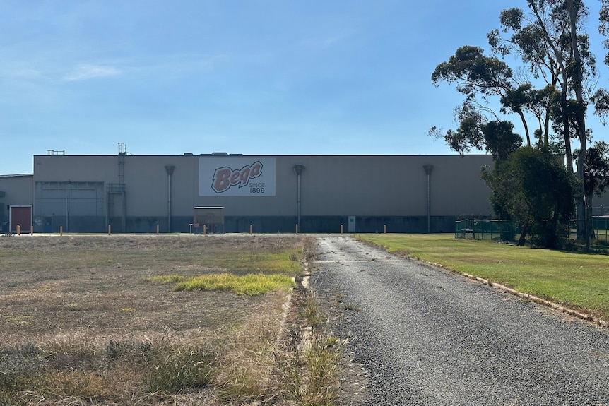 A large factory stand beside an empty grass field under a sunny sky.