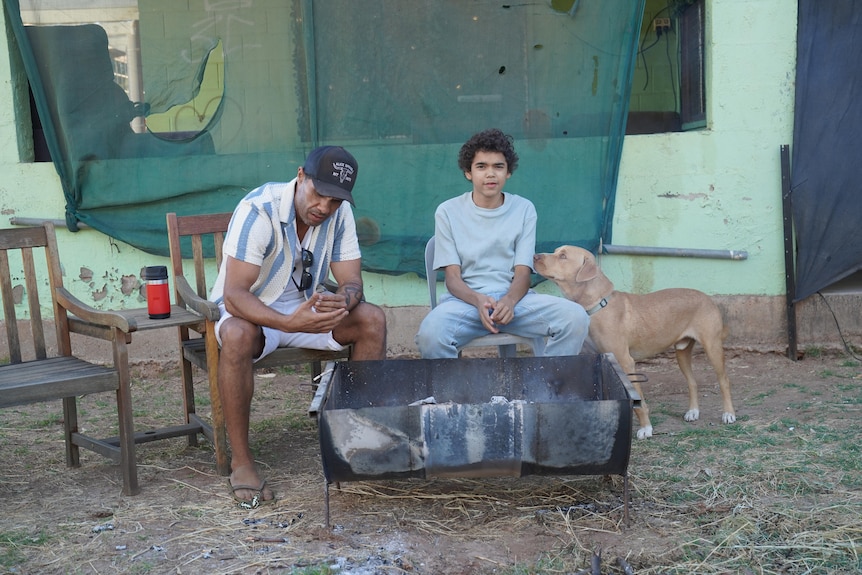 A man in a baseball cap, a young man and a dog sit beside a fire.