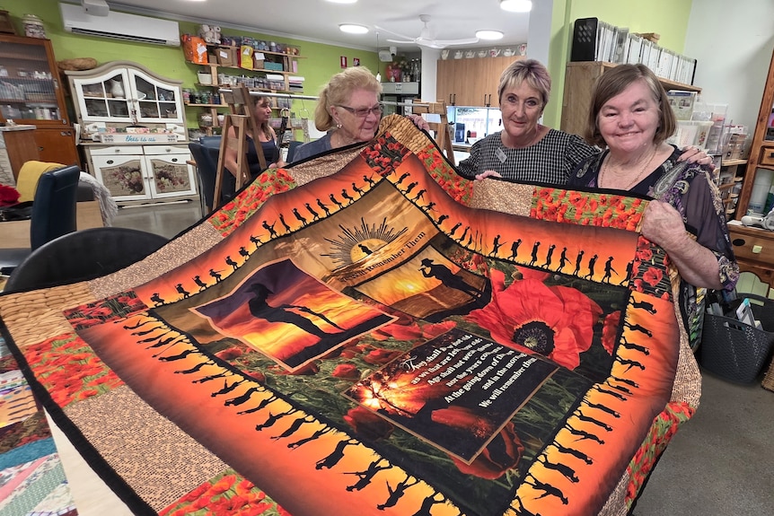 Three ladies hold up a bright orange handmade quilt.