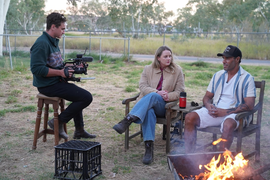 A young woman speaks to an Aboriginal man with a baseball cap as they sit beside a fire, while another man films them.