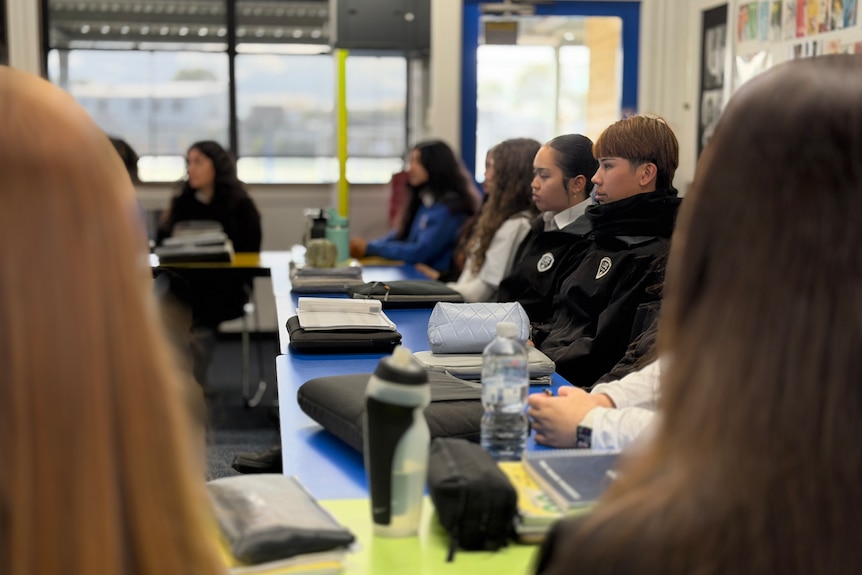Students sit at their desks looking ahead at the teacher.