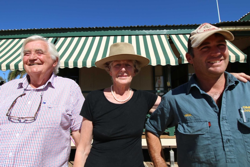 An older man and woman stand outside an outback home with a younger man.