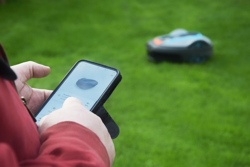 A man using a smartphone with a robot mower in the background