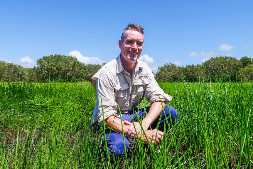 Matt crouches amid reeds, smiling at camera, blue skies, trees in the background.