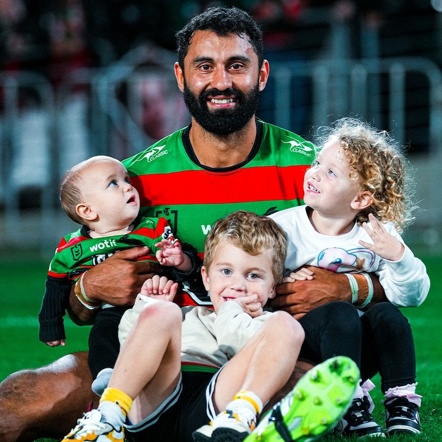 A Papua New Guinean man with a beard wearing a South Sydney Rabbitohs jersey smiles at the camera while holding 3 children 