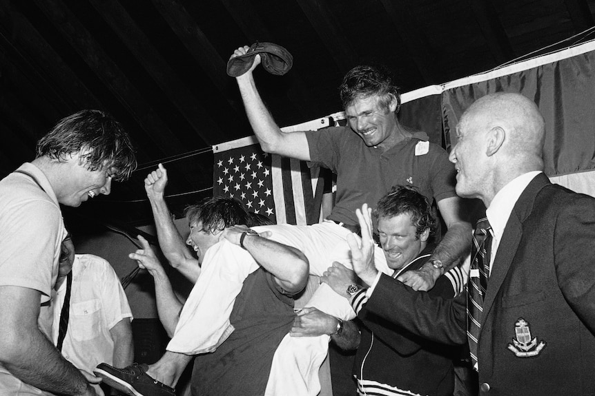 Ted Turner smiling and raising his arm in celebration being carried by celebrating yachtsmen.