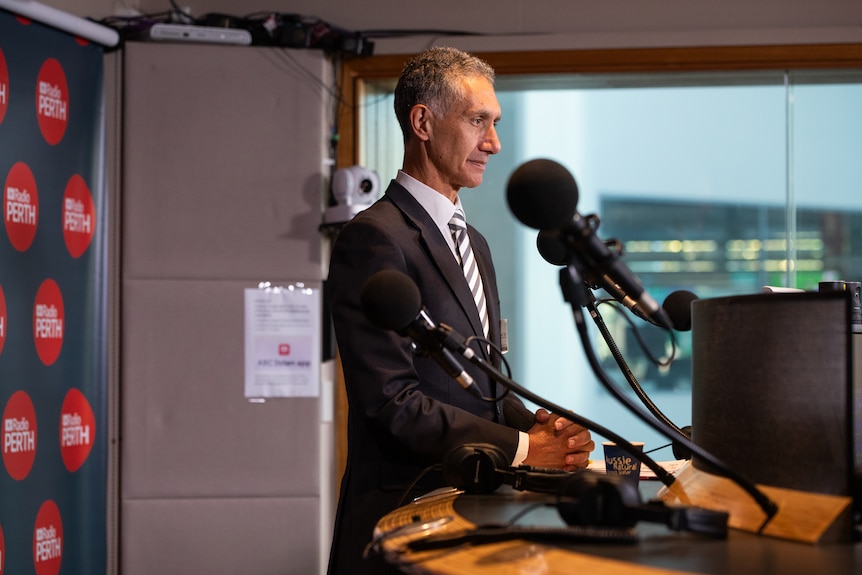 Tony sits at a round radio booth, microphones in front, is serious, wears suit, tie, ABC Perth signs on the wall behind.