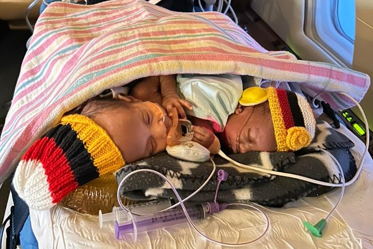 Twin babies sleep next to eachother underneath a blanket on a bed in a plane.