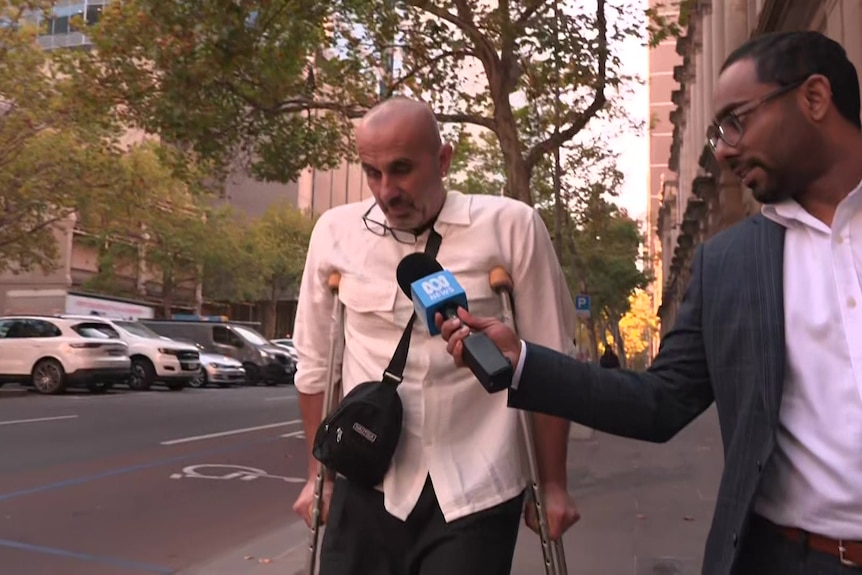 A dark-haired man in a suit holds a microphone up to a middle-aged man on crutches on a city street.