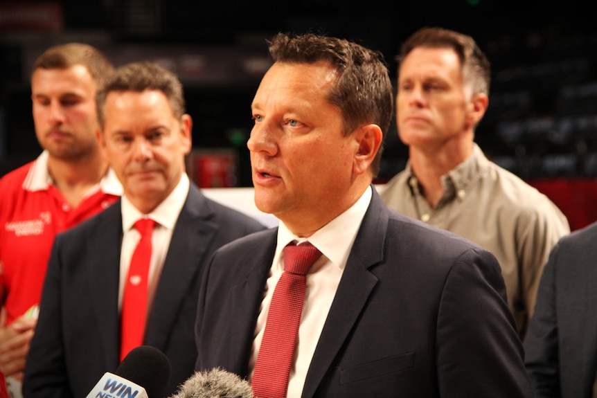 A dark-haired man in a suit speaks to the media while several other men watch on.