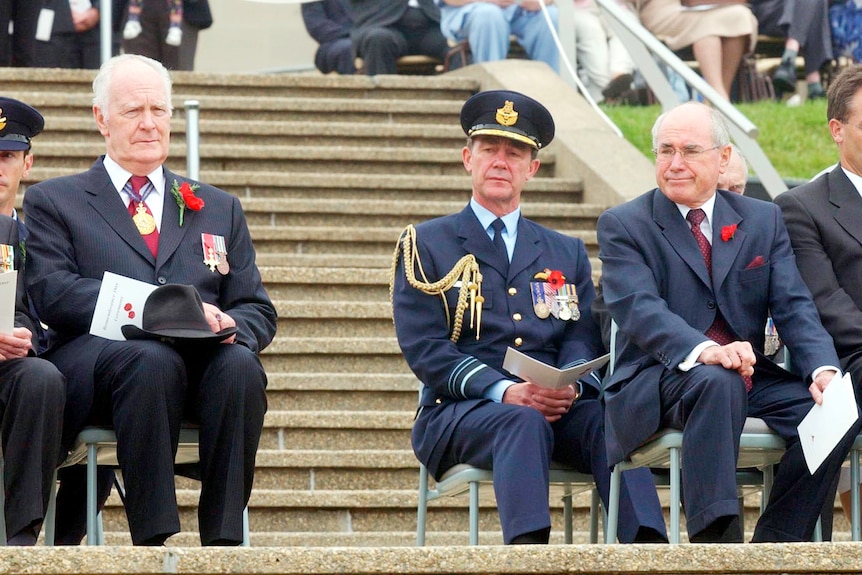 Three men in suits with military adornment sitting on chairs on steps
