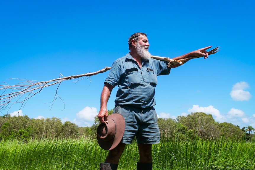 Christopher stands with a large branch over his shoulders, looking into the distance.