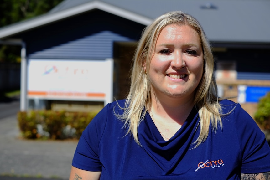 A woman with blonde hair in a blue shirt stands outside a building in the sunshine