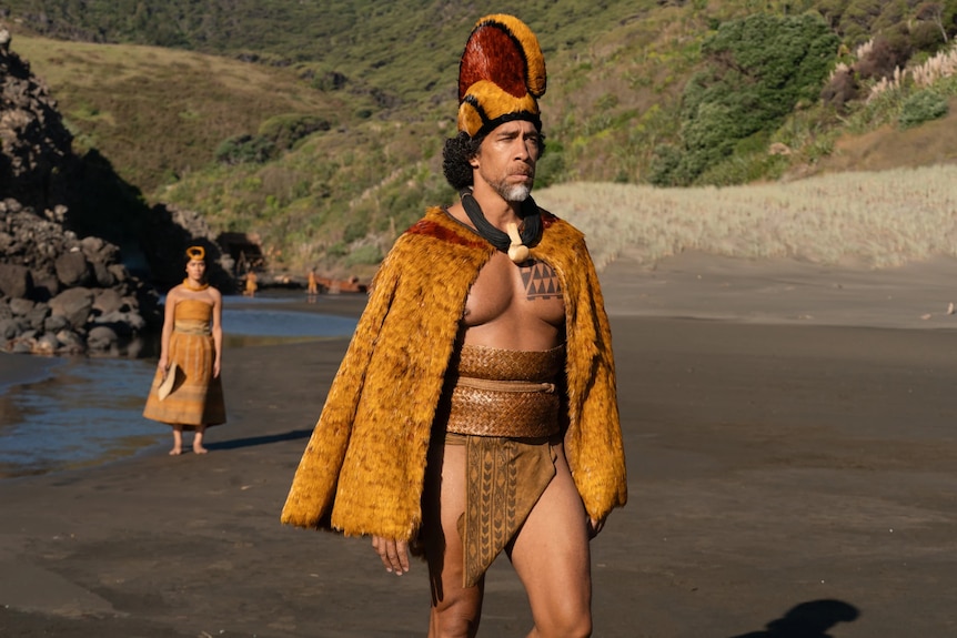 A man in a yellow and red outfit walks along a beach with a woman walking behind him