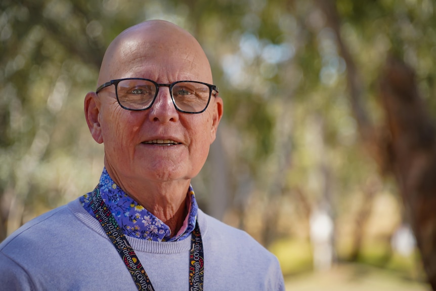 A photo showing a man wearing black glasses and blue printed shirt and light grey crewneck sweater