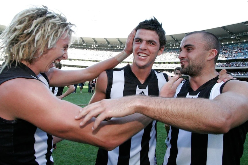 Three smiling AFL players celebrate while standing in a stadium.