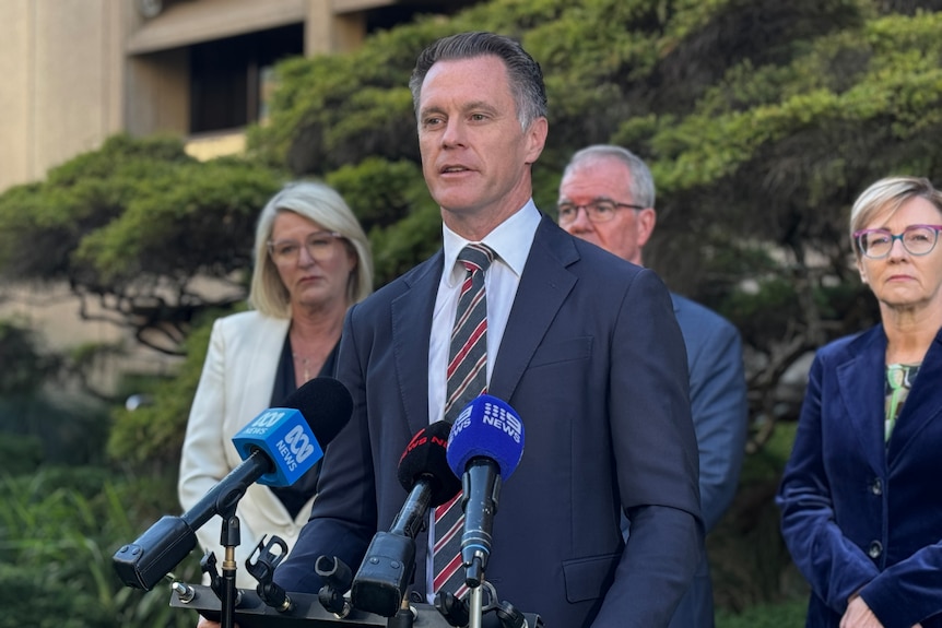 Man in charcoal suit speaks at press conference with a man and two women behind him in garden