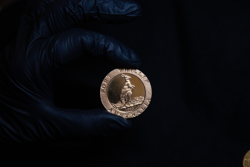 A person wearing a black rubber glove holds up an 1853 Port Phillip Kangaroo Office gold coin