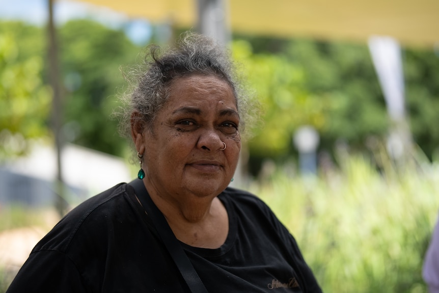 A middle aged Aboriginal woman with her greying hair tied back wearing a black tshirt sitting in a library's garden.