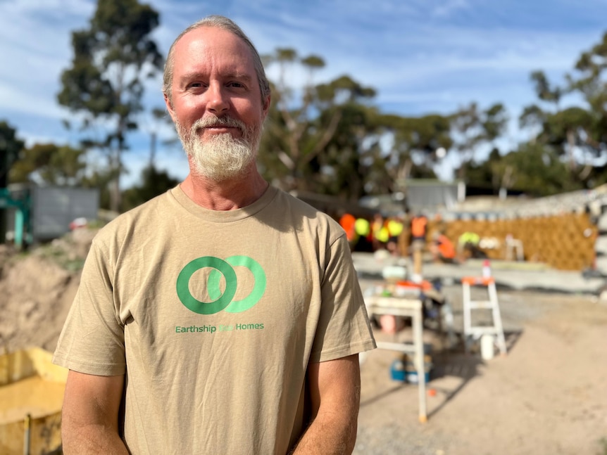 A man standing in front of an earthship smiling wearing a brown shirt