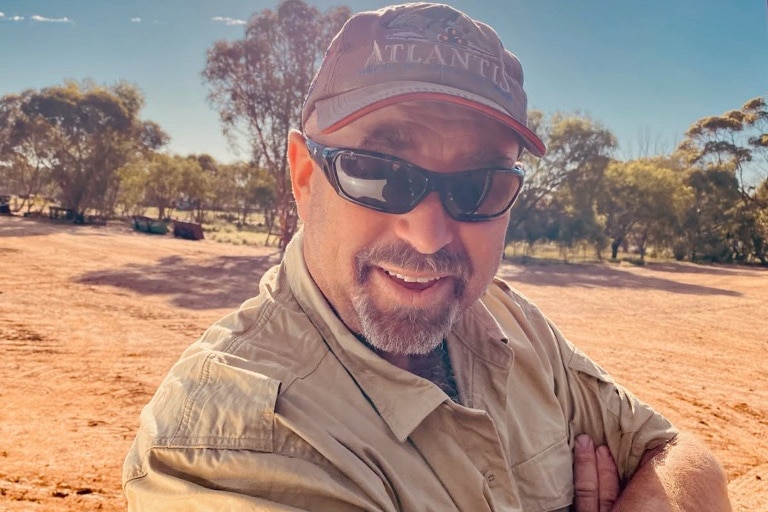 A man in sunglasses, a cap with a beard, smiling infront of a farm landscape. 