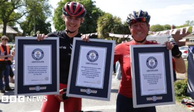 Josh and George Kohler are standing next to each other. They are both wearing red and black cycling wear and helmets. They are holding up three Guinness World Record certificates.