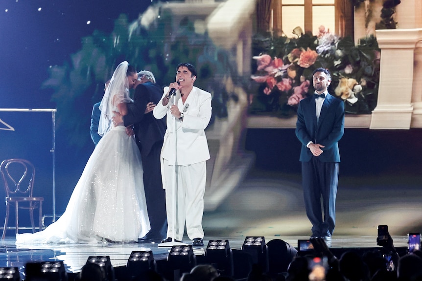Da Vinci, wearing a white suit, sings as behind him, a bride and groom look at one another