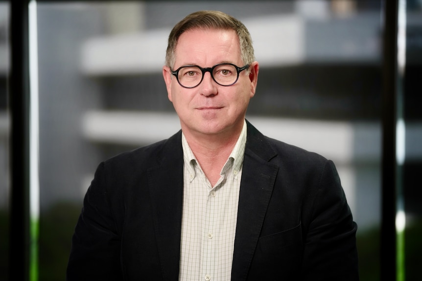 A man in a white suit and black jacket wearing thick framed glasses.