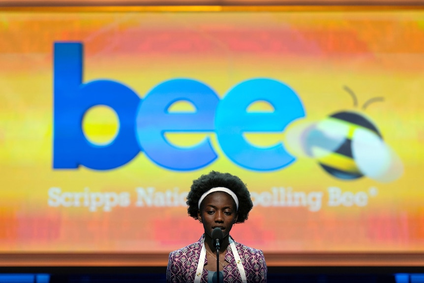 A girl stands on stage in front of a screen displaying the word BEE