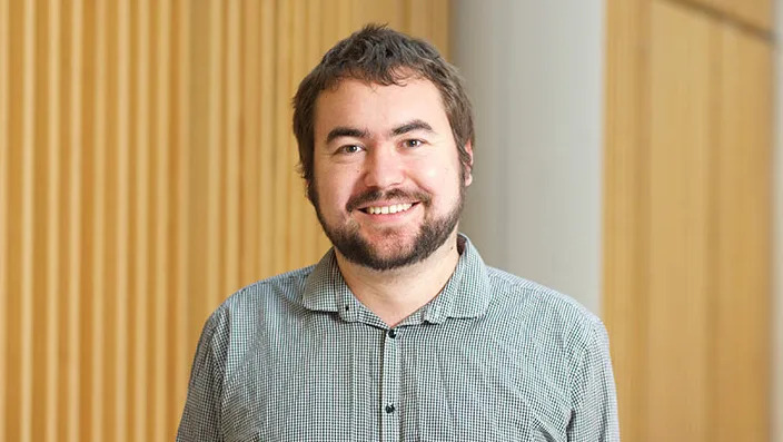 Dr Theo Hughes‑Riley has brown hair and beard, standing in a grey shirt