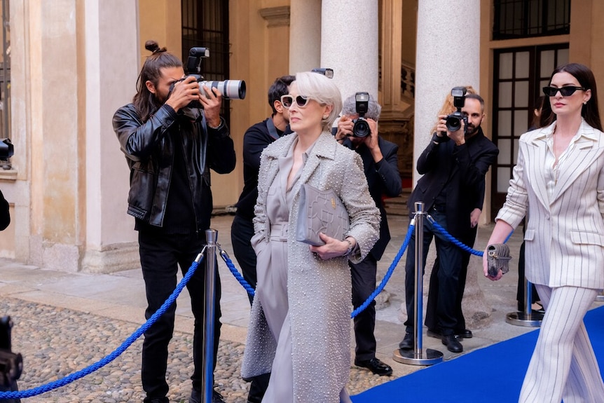Two women walk out in front of a pack on photographers