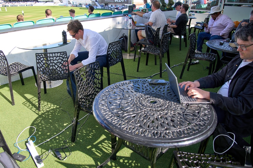 Men at laptops in an outdoor working area at a cricket ground.