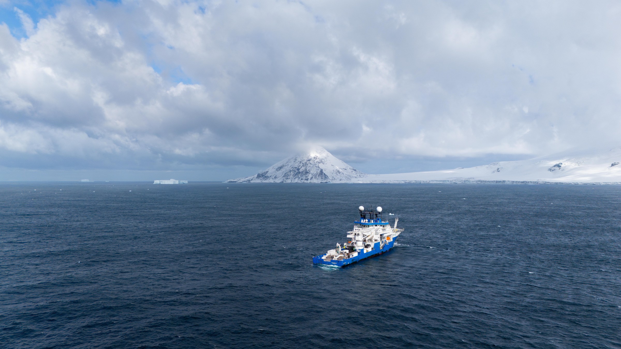 A research vessel, with a blue hull and lots of white research equipment on deck, sails on a dark sea with snow-covered shores and mountains in the background.