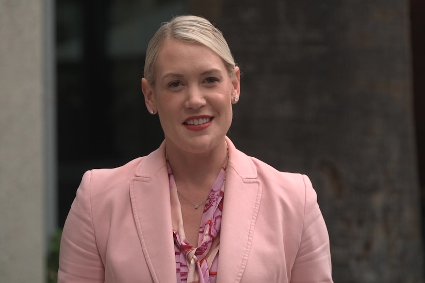 Ashton Hurn is wearing a pink jacket and speaking at a press conference