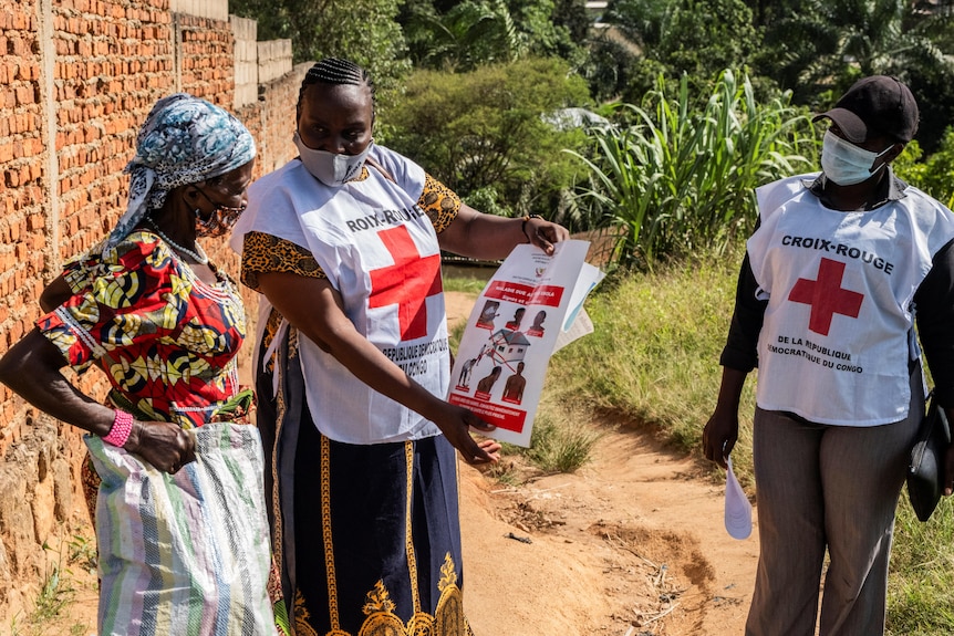 Two Red Cross employees wearing uniforms show a woman posters about the Ebola virus outside a house.