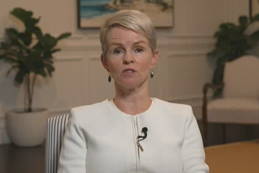 A blonde woman in a white jacket looks at the camera.