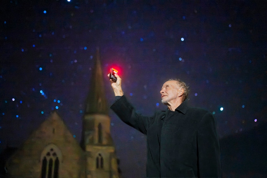 A gentleman in a black coat raises a device emitting a red glow into the air under a starry sky with church in background.
