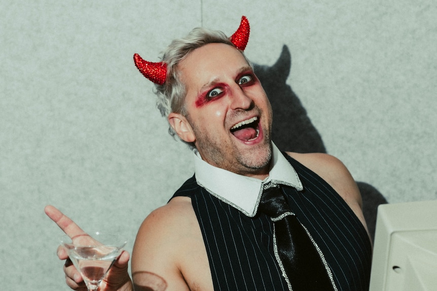 A comedian smiling, holding a drink and wearing devil horns.