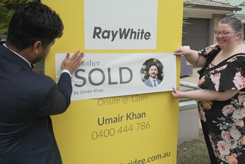 A woman and man place a sold sticker over a sale sign.
