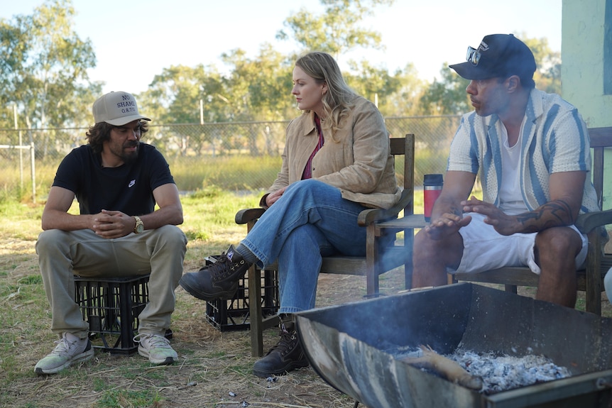 Two young men and a young woman talk as they sit near a fire.